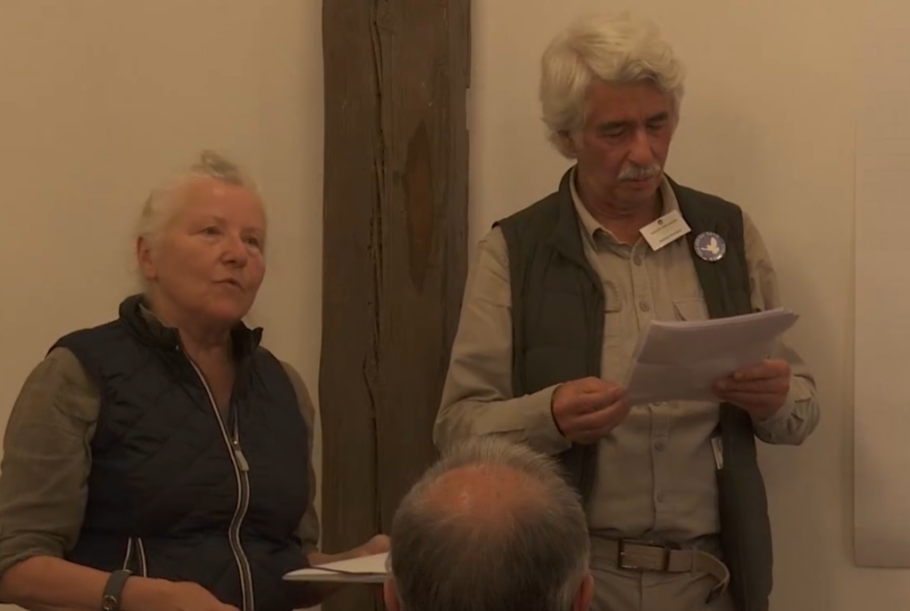 Fikentscher und Neumann beim Kongress 2025. Fikentscher steht links, trägt ihr Haar zurückgebunden und eine schwarze Weste. Sie hält Papiere in der Hand. Rechts von ihr steht Neumann, ebenfalls mit schwarzer Weste und Papieren in der Hand. Er trägt einen Button mit einer Friedenstaube.