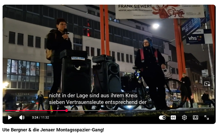 Ein Screenshot von YouTube, auf dem Bergner auf dem Holzmarkt in Jena links im Bild in ein Mikrofon spricht, dass an eine Anlage angeschlossen ist. Auf der rechten Seite lehnt Göring an seinem Fahrrad und sieht Bergner an. Der Titel des Videos lautet: "Ute Bergner & die Jenaer Montagsspazier-Gang!".