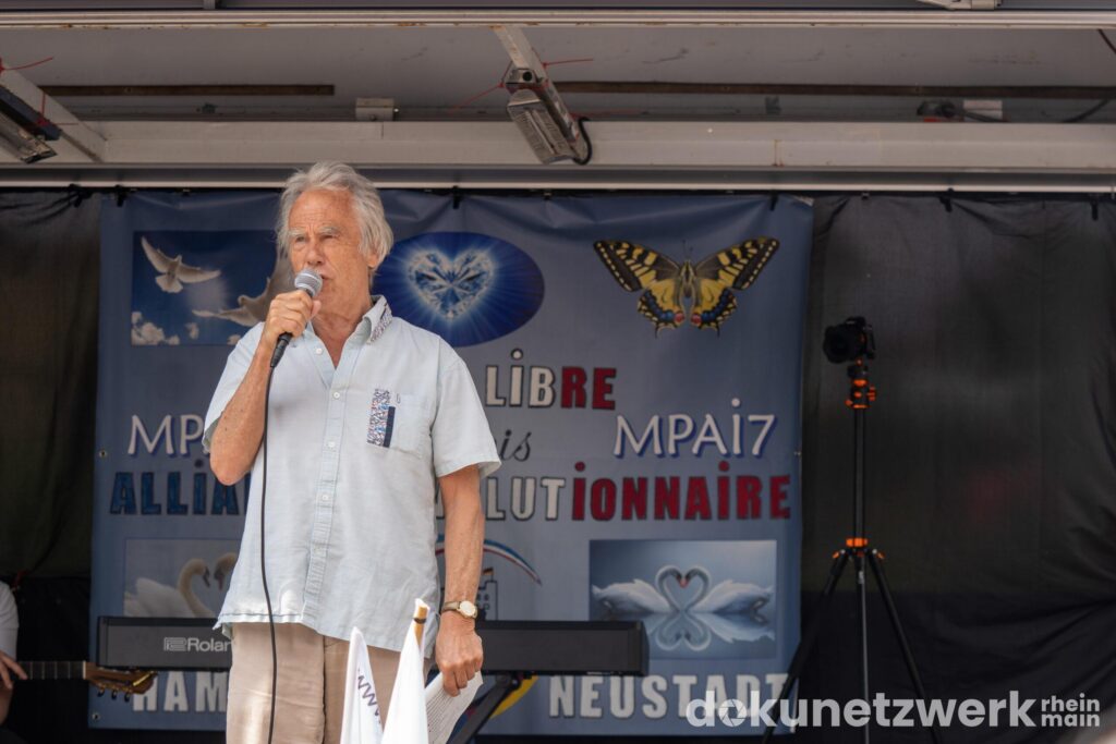 Reiner Braun steht auf einer Bühne, trägt ein helles Hemd und spricht in ein Mikrofon. Hinter ihm steht ein Keyboard und jemand hält eine Gitarre. Im Hintergrund hängt ein Transparen, auf dem in verschiedenen Sprachen das Wort "Frieden" gedruckt ist.