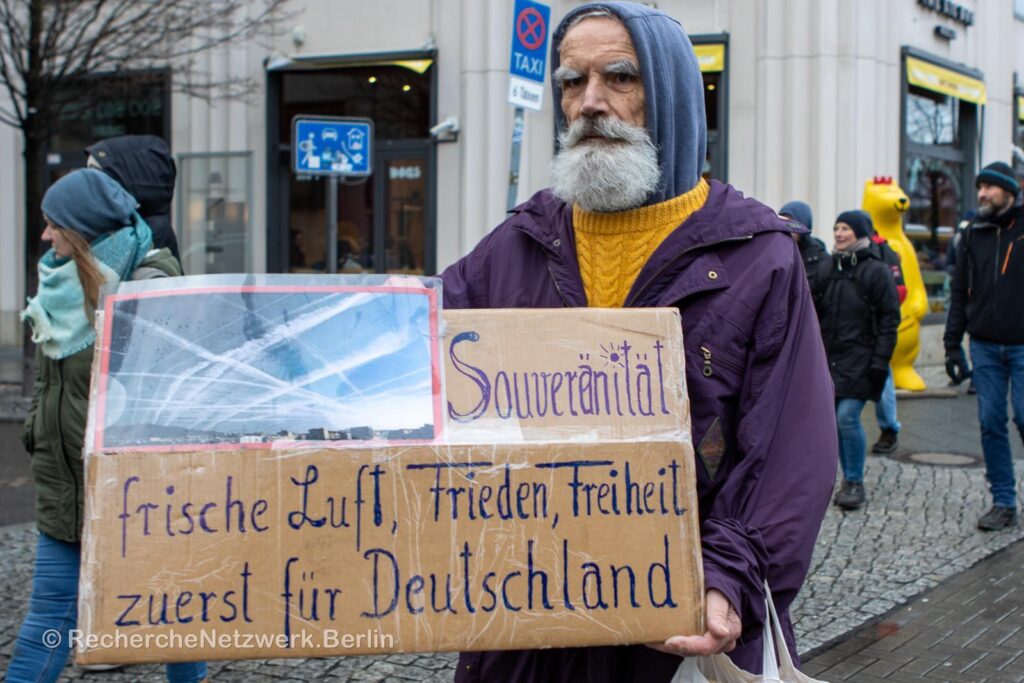 Sesterheim steht in einer Fußgängerzone und trägt einen gelben Pullover, eine lila Jacke und einen grauen Bart. Er hält ein Pappschild, auf dem steht: "Souveränität, frische Luft, Frieden, Freiheit, zuerst für Deutschland."