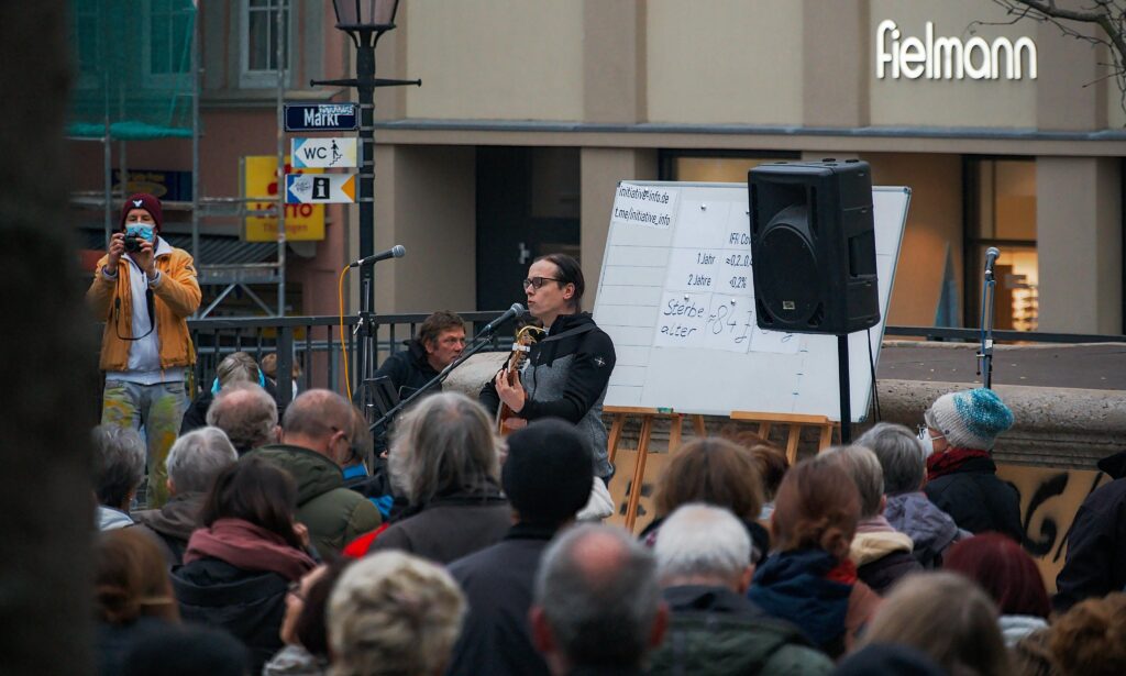 Jan Wetzel spielt Gitarre und singt in eine Mikrofon. Im Vordergrund sieht man die Teilnehmenden der Demo, links im Bild steht ein Fotograf in gelber Jacke und mit medzinischem Mundschutz. Im hIntergrund sieht man eine weiße Tafel mit unleserlicher Schfrit und eine Fielmann-Filiale.